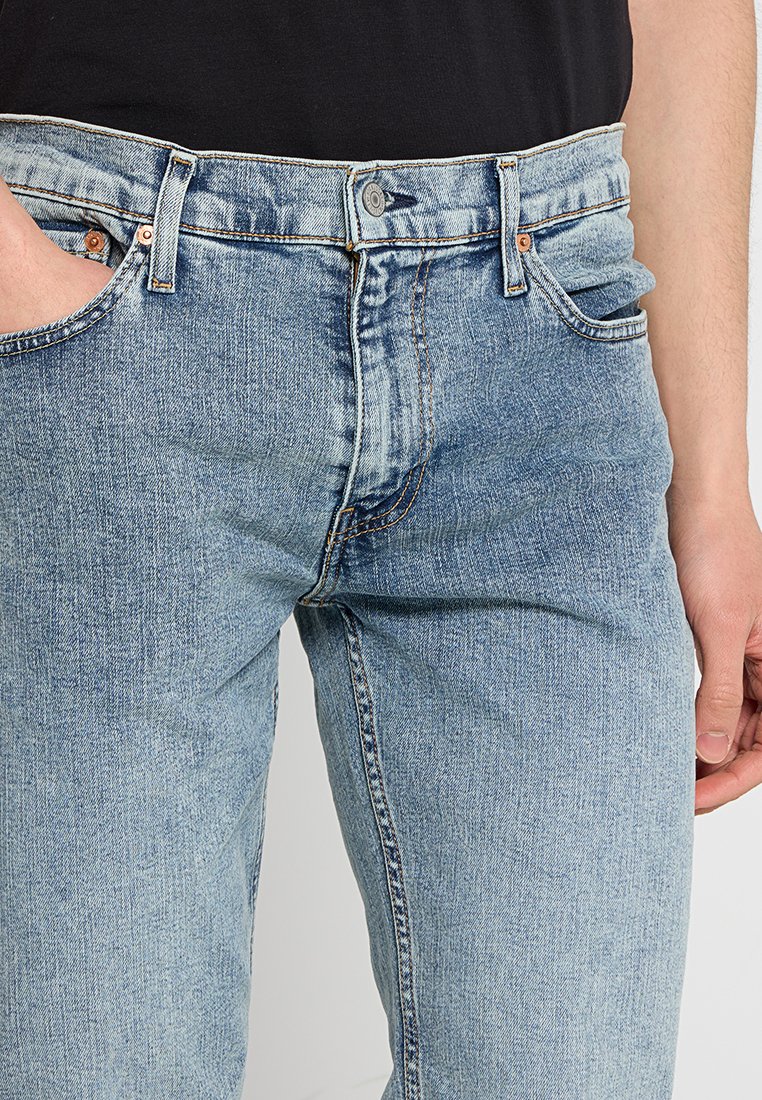 Los jeans de mezclilla azul claro presentan un diseño clásico de cinco bolsillos, cierre de botón y un ligero desgastado para un aspecto usado. Textura suave con acentos de costura.
