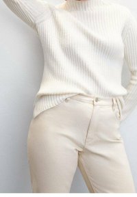 Suéter de punto acanalado color crema con un cuello alto, combinado con jeans de color beige claro, que cuentan con un bolsillo frontal y un cierre de cremallera.