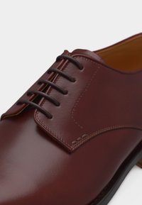 Κοντινή λήψη ενός γυαλισμένου καφέ δερμάτινου παπουτσιού για επίσημη ενδυμασία με τέσσερις οπές και σκούρα καφέ κορδόνια σε λευκό φόντο.