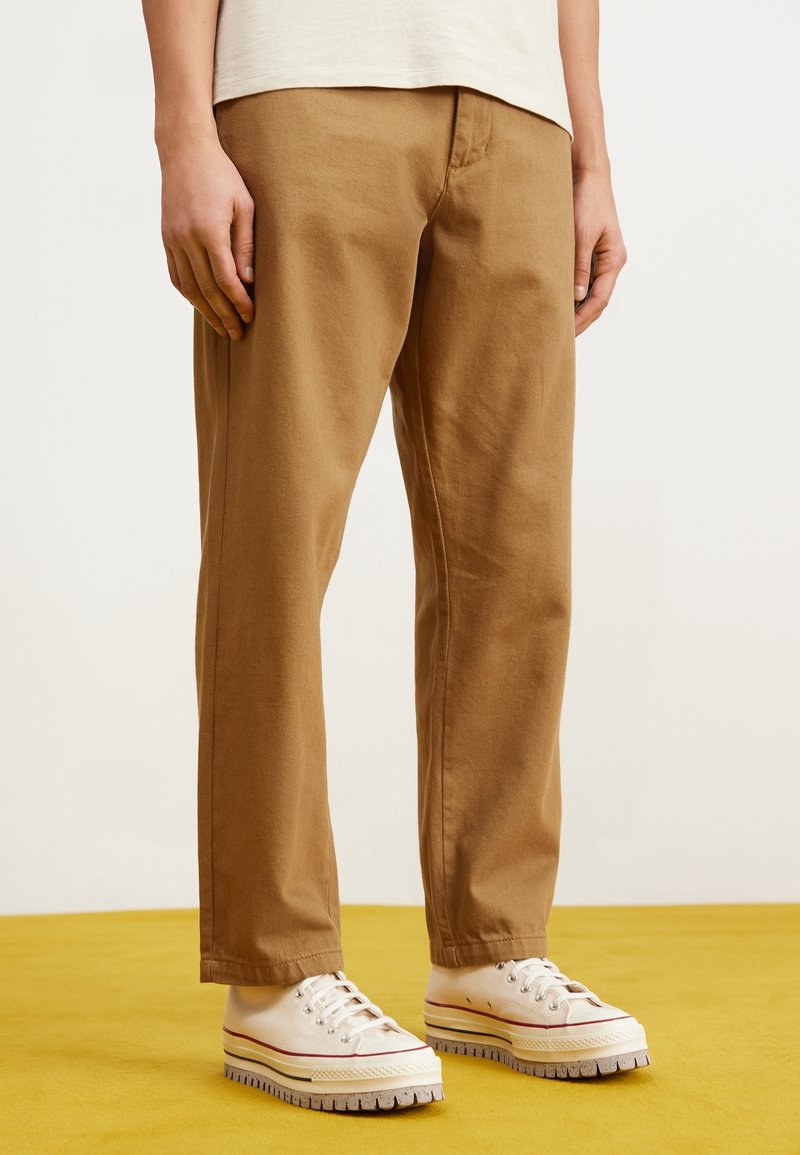 Pantalon en coton marron à coupe décontractée, jambes droites, avec des détails de coutures visibles, associé à des baskets blanches avec des accents colorés.