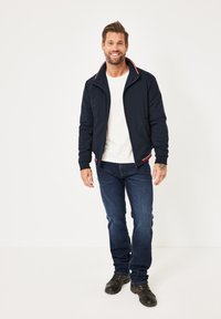 Mann lächelt in einer marineblauen Reißverschlussjacke mit rot-weißer Borte, weißem T-Shirt, blauen Jeans und schwarzen Stiefeln, vor einem weißen Hintergrund stehend.