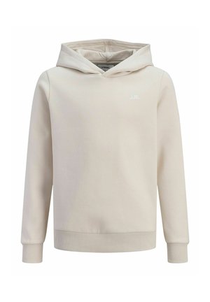Garçon debout les mains dans les poches, portant un sweat à capuche beige, un pantalon de survêtement assorti et des baskets blanches, regardant sur le côté devant un fond uni.