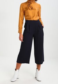 Kulta samettitoppi, jossa on solmukkaat kaula-aukko, yhdistettynä tummansinisiin leveälahkeisiin housuihin; malli käyttää valkoisia nilkkureita.