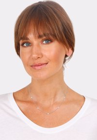 Silver necklace with five small square accents, delicate chain, worn over a white shirt with a wide neckline. Smooth texture and polished finish.