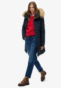 Mørkeblå dunjakke med en hætte med pelskant, rød sweater, blå jeans og brune støvler med gule snørebånd. Med lynlåslommer.