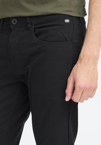 Jean en denim noir présentant un design classique à cinq poches, une fermeture à bouton et une petite étiquette de marque sur la poche avant. Texture lisse.
