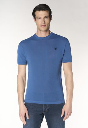 Uomo che indossa una maglietta blu a maniche corte con scollo tondo e un piccolo logo nero ricamato sul petto sinistro, abbinata a jeans blu scuro.