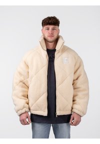 Chaqueta de felpa color crema con un diseño de rombos acolchados. Cuello alto, cremallera frontal y parche de logo en el lado izquierdo del pecho. Textura suave.