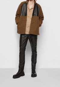 Brun fleecejacka med svarta detaljer i konstläder, ihop med svarta läderbyxor och svarta snörboots. Dragkedja framtill.