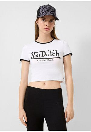 Weißer Baumwoll-Crop-Top mit schwarzem Saum, der "Von Dutch ORIGINALS" in fettem schwarzen Text bedruckt hat. Kombiniert mit schwarzen Leggings.