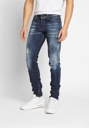 Vaqueros ajustados de mezclilla azul con detalles desgastados y desvanecimiento en azul claro, combinados con zapatillas blancas.