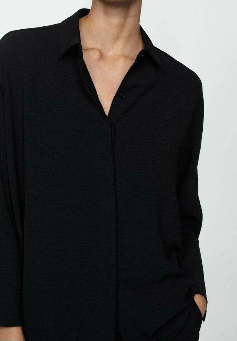 Chemise noire à manches longues avec boutons et col, en tissu légèrement texturé et coupe décontractée, poche avant sur le côté gauche.