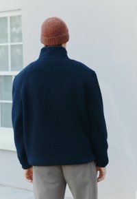 Person iført en brun strikket beanie, en marineblå fleecejakke og grå bukser, der står med ansigtet mod en hvid væg med et vindue til venstre.