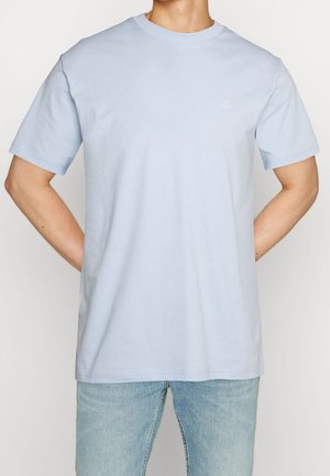 Mężczyzna w jasnoniebieskim t-shircie z krótkim rękawem z małym białym logo na piersi i jasnoniebieskich dżinsach, ręce umieszczone za plecami.