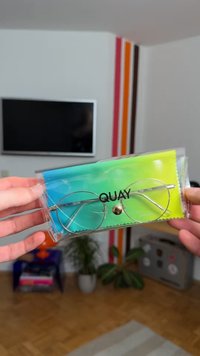 Sølvmetal briller med runde linser, i en gennemsigtig emballage med en gradient blå og grøn renseklud. Indeholder "QUAY" branding.
