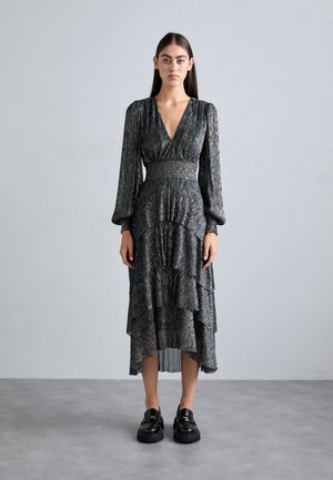 Robe midi plissée noire avec des manches longues, un décolleté en V prononcé et un ourlet en tiers. Fabriquée dans un tissu texturé brillant, associée à des chaussures noires épaisses.