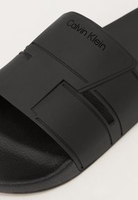 Sandalo nero a infradito con soletta in gomma testurizzata e una fascia ampia. Presenta il logo "Calvin Klein" in rilievo e motivi geometrici sulla fascia.