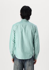 Camicia verde chiaro con bottoni, realizzata in un tessuto di trama. Presenta maniche lunghe e un orlo curvo con polsini abbottonati.