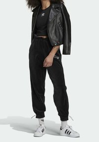 Svart läderbikerjacka över en svart kortärmad t-shirt, parat med svarta velourjoggingbyxor med vita logodetaljer och vita Adidas-sneakers.