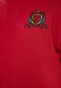 Felpa in cotone rossa con un ricamo che presenta uno stemma con una corona, stelle e un nastro con la scritta "dal 1998". Texture liscia e vestibilità casual.