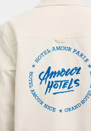 Veste écrue avec texte brodé bleu au centre indiquant « Amour Hotels » et entouré des inscriptions « Hotel Amour Paris », « Hotel Amour Nice » et « Grand Hotel ».