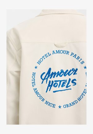 Kreminės spalvos striukė su mėlynu siuvinėtu užrašu centre: „Amour Hotels“, o aplink - „Hotel Amour Paris“, „Hotel Amour Nice“ ir „Grand Hotel“.