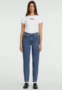 Bela kratka majica s kratkimi rokavi z rdečim logotipom "GUESS", v kombinaciji z oprijetimi modrimi jeans hlačami. Model nosi črne mokasinke. Preprost ozadje.