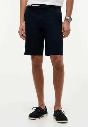 Man draagt marineblauwe shorts, zwarte veterschoenen, een wit overhemd in de broek gestopt, een zwarte riem met rode en witte strepen en een polshorloge.