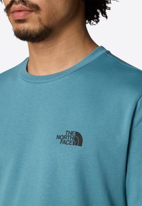 Camiseta de algodón azul con escote redondo, que presenta un logo negro de "The North Face" en el área del pecho izquierdo. Textura suave.