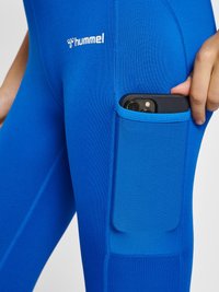 Blå leggings med en texturerad ficka, som har en telefon. Materialet är elastiskt, designat för komfort och bekvämlighet, med en logotyp på låret.