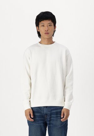 Vit sweatshirt med rund halsringning, långa ärmar och ribbade cuffar. Kombinerad med blå denimjeans, uppställd mot en enfärgad bakgrund.
