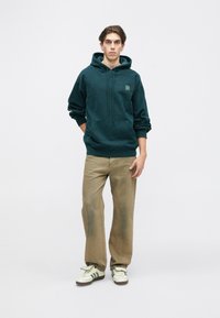 Hoodie en polaire vert foncé avec une poche kangourou, associé à un jean beige ample et des baskets blanches avec des rayures noires. Le mannequin se tient droit.