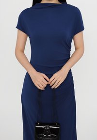 Mörkblå, kortärmad, draperad klänning med draperingsdetalj vid midjan, tillsammans med en liten svart axelväska med en dekorativ lås.