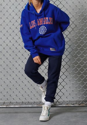 Blå hoodie med "LOS ANGELES" i röd text, matchad med marinblå byxor och vita sneakers med gröna detaljer. Mjuk, avslappnad material.