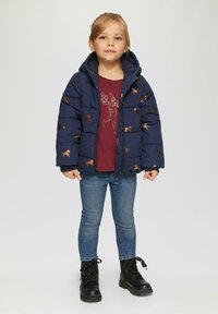 Doudoune marine avec des motifs de chevaux dorés, design à capuche, associée à une chemise bordeaux, un jean bleu et des bottes noires à lacets.