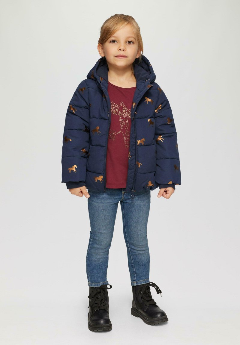 Doudoune marine avec des motifs de chevaux dorés, design à capuche, associée à une chemise bordeaux, un jean bleu et des bottes noires à lacets.