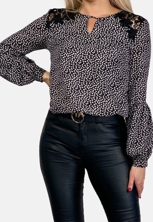 Kvinde iført en sort og hvid langærmet bluse med pletter og blonde detaljer på skuldrene samt sorte læderbukser med bælte.