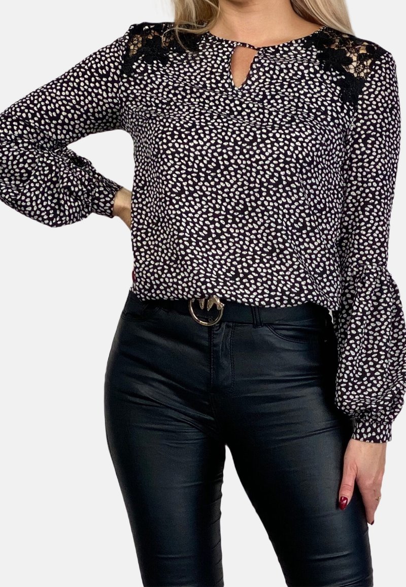 Femme portant un chemisier à manches longues noir et blanc à pois avec des détails en dentelle sur les épaules et un pantalon en cuir noir avec une ceinture.