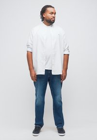 Άνδρας με πλεγμένα μαλλιά που φοράει λευκό πουκάμισο με κουμπιά πάνω από λευκό μπλουζάκι, μπλε τζιν και μαύρα αθλητικά παπούτσια, στέκεται μπροστά σε απλό φόντο.