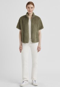Kurzärmlige olivgrüne flauschige Jacke mit hohem Kragen, kombiniert mit einem hellen Oberteil und weißen Hosen, mit vorderen aufgesetzten Taschen und Sneakers.