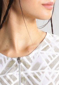 Biała bluzka zapinana na zamek z geometrycznymi złotymi i jasnoszarymi wzorami, teksturowany materiał, okrągły dekolt i krótkie rękawy. Widoczny detal zamka.