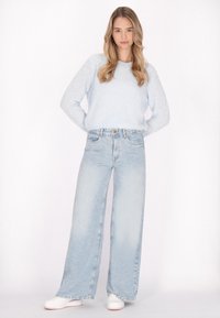 Heller blauer, flauschiger Pullover mit langen Ärmeln, kombiniert mit hoch taillierten, weit geschnittenen hellblauen Jeans und weißen Sneakers. Schlichte Accessoires.