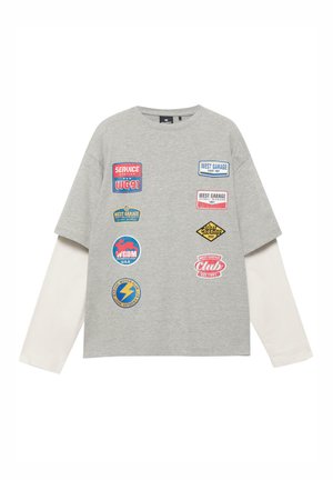 Maglia grigia a maniche lunghe a strati con maniche bianche, con nove patch colorate in stile vintage di garage e club motoristici sul davanti.