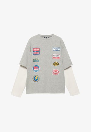 Maglia grigia a maniche lunghe a strati con maniche bianche, con nove patch colorate in stile vintage di garage e club motoristici sul davanti.