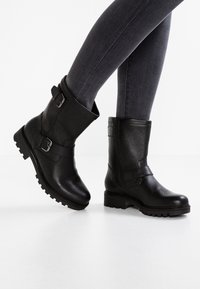 Botines de cuero negro hasta el tobillo con hebillas, acabado texturizado y suelas resistentes, combinados con jeans ajustados gris oscuro sobre una superficie blanca.