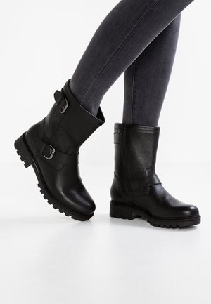 Botines de cuero negro hasta el tobillo con hebillas, acabado texturizado y suelas resistentes, combinados con jeans ajustados gris oscuro sobre una superficie blanca.