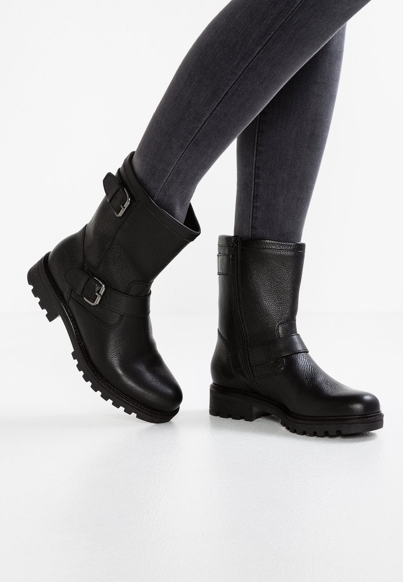 Botines de cuero negro hasta el tobillo con hebillas, acabado texturizado y suelas resistentes, combinados con jeans ajustados gris oscuro sobre una superficie blanca.