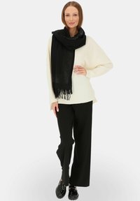 Czarny szalik z frędzlami, z subtelnym wzorem kropek, w połączeniu z białym, oversize'owym swetrem i czarnymi spodniami typu wide-leg. Błyszczące czarne buty.