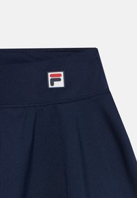 Jupe de sport bleu marine avec une taille élastique, arborant un petit logo Fila rectangulaire en rouge, blanc et noir au niveau de la taille. Texture douce.