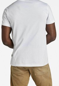 Camiseta blanca de algodón con mangas cortas y cuello redondo, vista desde la parte de atrás. Diseño simple, sin patrones ni gráficos visibles.